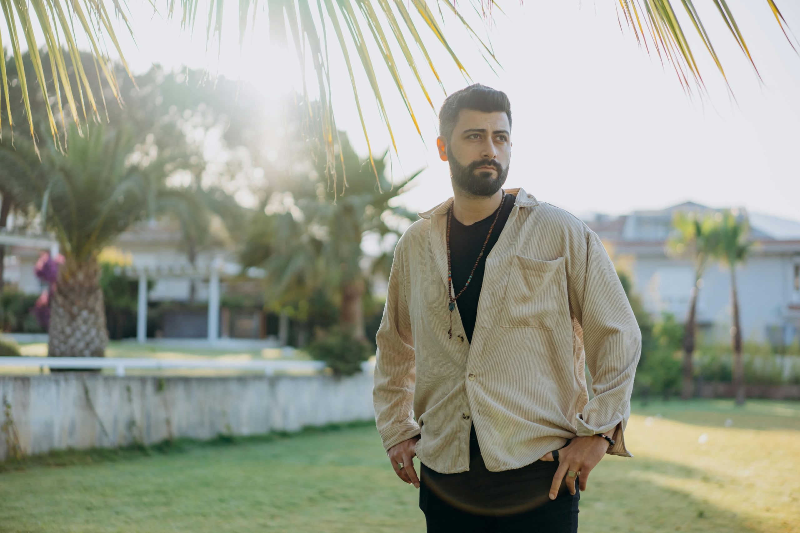 Fashionable man with beard posing confidently outdoors in a sunlit garden, showcasing style and elegance.
